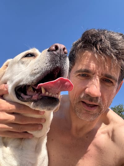 A fair-skinned man with short gray hair, a mustache, and light-colored eyes, is holding a light-colored dog with its mouth open and tongue protruding. The man's right arm is visible, cradling the dog. The background is a clear blue sky. The photograph appears to have been taken at a low angle, looking up at the subjects. The location is not discernible as being specifically in Kato Polemidia Municipality, Cyprus based on the image provided.
