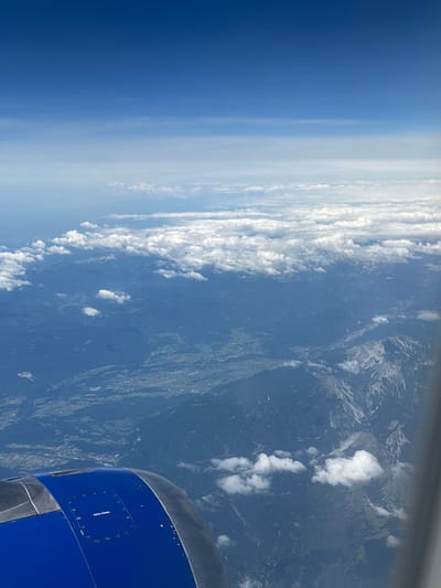 An aerial view captures the mountainous landscape surrounding Viehhausen, Austria, seen from an airplane window. In the foreground, the blue and gray engine cowling of the aircraft is visible. Below, rugged, dark green mountains stretch towards the horizon, their peaks partially obscured by fluffy white clouds. Patches of lighter green indicate valleys and possibly agricultural land, with a small, shimmering body of water, likely a lake, nestled within the terrain. The sky above is a gradient of deep blue, transitioning to lighter shades as it meets a haze of clouds in the distance. The overall scene depicts a serene, natural environment viewed from high altitude.