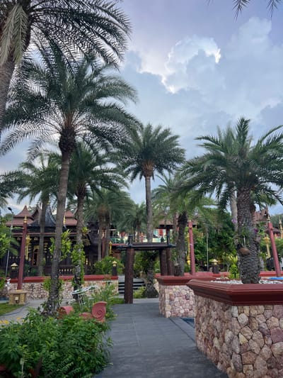 The image depicts a serene courtyard or garden area in Baan Lamai, Thailand, likely a resort or temple. Tall palm trees dominate the foreground, their fronds silhouetted against a partly cloudy sky. In the background, a traditional Thai-style building with intricate dark wood and gold accents is visible, featuring multiple tiered roofs. The building is flanked by lush green vegetation. Stone walls with decorative red trim frame a pathway leading into the scene. The overall atmosphere is tranquil and tropical, suggesting an outdoor recreational or cultural space. There are no people visible in the image.