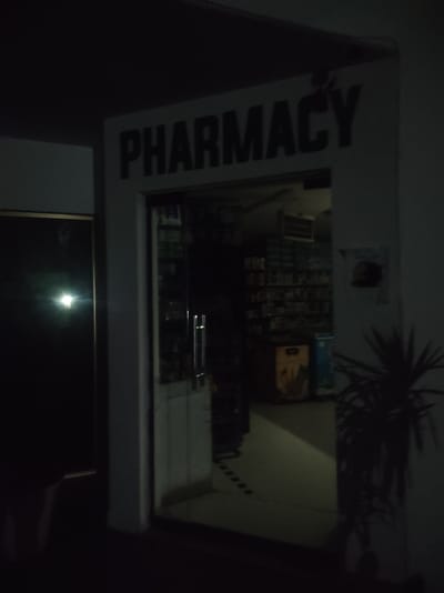 The image shows the entrance to a pharmacy at night. The word "PHARMACY" is prominently displayed in large black letters on a white sign above the entrance. The doorway itself is open, revealing a dimly lit interior with shelves stocked with products. To the left of the entrance is a dark, reflective surface, possibly a window or a door. To the right of the entrance, a potted plant with spiky leaves casts a shadow on the wall. The overall environment suggests a commercial establishment in Roranwala, Pakistan, operating during nighttime hours.