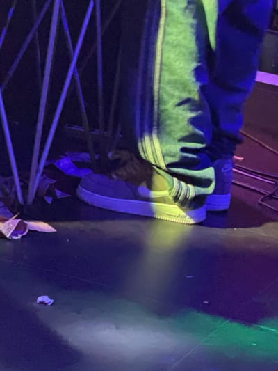 A person wearing gray Nike Air Force 1 sneakers and a light green and black jacket is standing behind a blue metal railing. Debris is scattered on the ground near the feet. No information about the location being in Obosi, Nigeria, is available in the image.