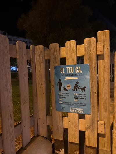 This media file shows a wooden structure, possibly a bench or fence, in an outdoor setting at night. A blue sign is attached to the wooden structure, featuring text and illustrations related to dog ownership. The sign, which appears to be in Catalan, states "EL TEU CA, LA TEUA RESPONSABILITAT" (Your dog, your responsibility). It includes illustrations depicting people with dogs, one with a dog excrement being picked up, and another showing a dog wearing a muzzle.

The text on the sign details obligations for dog owners, including the requirement to keep dogs leashed ("FERMATS") and to pick up their excrement ("RECOLLIR ELS SEUS EXCREMENTS"). It also mentions that all cataloged dogs that are potentially dangerous must wear a muzzle ("HAN DE DUR MORRAL"). A penalty of up to 1500€ is indicated for non-compliance ("NO FER-HO PORTA SANCIONS FINS A 1500 €"). The sign also includes the logo of "d'eivissa," suggesting the location is Ibiza, Spain.

The scene is illuminated by artificial light, casting a warm glow on the wooden structure and the sign. The background is dark, with the faint outline of trees and a building visible, indicating a nighttime setting in what appears to be an urban or park area. The overall mood is informative and regulatory, emphasizing the responsibilities of pet owners in a public space.