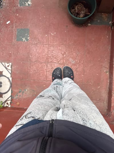 An overhead shot shows a person standing on a red tile floor wearing black Crocs and gray patterned fleece pants, along with a dark navy blue jacket. To the left, a green bucket contains dried plant matter. The floor appears to be indoors or on a porch. Below the Crocs, the tiles are decorative with a black and white pattern. The location is presumed to be Avellaneda, Argentina.