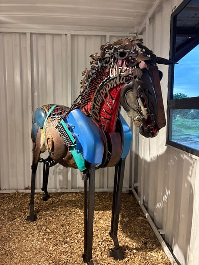 In the city of Karum, Nigeria, a large, intricately constructed metal sculpture of a horse stands indoors, possibly in a workshop or gallery. The sculpture is made from a variety of repurposed metal parts, including what appear to be car parts, chains, and other industrial scraps, assembled to form the horse's body, legs, and head. The horse has a distinctive mane and tail fashioned from metal fragments. Brightly colored plastic elements, such as blue and green pieces, are incorporated into the sculpture, particularly around the neck and saddle area. A red material with white lettering, possibly a sign or fabric, is visible within the horse's neck. The sculpture is supported by dark metal legs and stands on a floor covered with wood chips or shavings. The background consists of corrugated metal walls painted white, with a white ceiling. To the right of the sculpture, a large window offers a view of an outdoor landscape with trees and a cloudy sky. The lighting suggests an indoor setting with some natural light entering from the window.