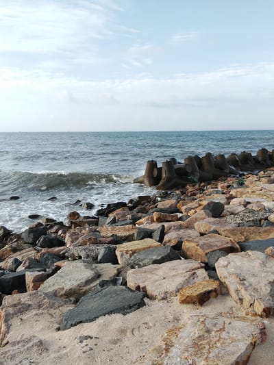 A wide shot captures a rocky shore with the ocean in the background. The sky is a pale blue with scattered clouds. The ocean is a deep blue-grey, with moderate waves breaking on the shore. The shore is covered in rocks of various sizes and colors, from light tan to dark grey. In the distance, a series of concrete tetrapods form a breakwater. The sand is a light tan color and appears damp in places. The overall scene is one of a rugged coastline.