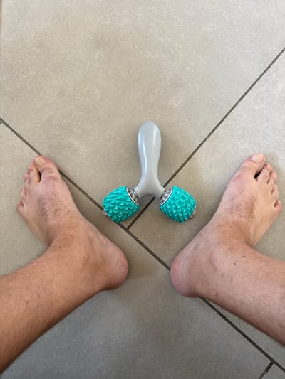 An overhead, wide shot captures a personal foot massager positioned between two bare feet on a light gray, tiled floor. The massager is a light gray handheld device with two turquoise, spiked rollers. The feet, one on the left and one on the right, are positioned facing upwards, with the toes slightly spread. The floor tiles are laid in a grid pattern, with dark gray grout lines. The lighting is bright and even, suggesting an indoor setting. No other individuals or objects are visible in the frame. The location of Drumettaz-Clarafond, France is irrelevant to the visual content of the image.