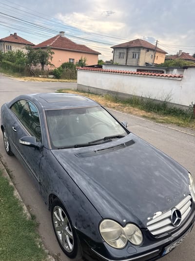 This is a media file featuring a dark-colored Mercedes-Benz car, possibly a CLS model, parked on the side of a paved road. The car appears to be dusty, with streaks and smudges visible on its surface. The perspective is from above and to the side, looking down at the car's hood, windshield, and side. The car's front headlights and grille, with the Mercedes-Benz logo, are visible. A sunroof is also visible on the roof.

The setting is a residential area, indicated by houses with tiled roofs visible in the background. One house is a light peach color, and another appears to be a lighter gray or beige. There are trees and vegetation surrounding the houses and along the roadside. Overhead power lines are also prominent in the scene, suggesting an urban or semi-urban environment.

The sky is overcast with scattered clouds, indicating a cloudy or partly cloudy day. The lighting suggests it might be late afternoon or early evening. There is no clear indication of any specific activity or event taking place; the car appears to be parked.

One license plate is partially visible on the front of the car, displaying some numbers and letters, including what appears to be "C 563..." and possibly Bulgarian characters. The overall impression is a snapshot of a car in a residential setting under a cloudy sky. The location context provided is Kostinbrod, Bulgaria.