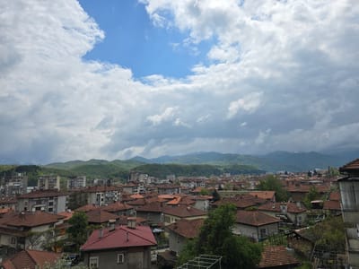 The image depicts an elevated view of a residential area in Troyan, Bulgaria, under a sky dominated by cumulus clouds with patches of blue. The landscape includes dense groupings of buildings, primarily houses with red-tiled roofs, interspersed with trees and greenery. In the distance, there's a range of forested hills. Some taller, multi-story apartment buildings are visible within the urban landscape, indicating a mix of residential building styles. The overall setting suggests a residential area on the outskirts of the town.