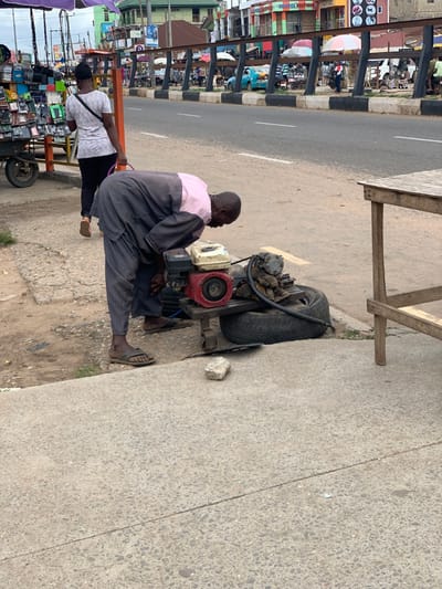 A man in a pink and grey shirt and grey pants is working on a small engine on a wheeled platform. He is crouched down and looking at the engine. The engine is red and white with a black handle. The platform has a large black tire attached to it. There is a small rock and a white plastic lid on the ground near the platform. A woman in a pink shirt and grey pants is walking away from the scene on the sidewalk. There is a wooden table to the right of the man. The scene is in Ondo, Nigeria, at latitude 7.252 and longitude 5.199. There is a busy road in the background. The buildings in the background are concrete. There is a black metal fence in the foreground.
