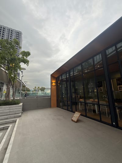 The image shows an exterior view of a modern cafe or restaurant named "CLOUD" located in Bangkok, Thailand. The cafe features large glass windows and a wooden facade, with an outdoor seating area paved with large gray tiles. A small, A-frame sign with text is placed on the ground in front of the cafe. To the left, a modern tall building stands against a cloudy sky. A tree with green foliage is planted in a planter box, and a person is seated on a ledge in the background. The overall scene suggests a contemporary urban environment.