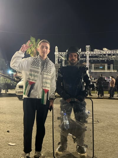 The image, taken at night in what is identified as Al-Muhajerin Municipality, Syria, features two men posing outdoors in what appears to be an event or public gathering space. The background is dark, illuminated by numerous artificial lights on large metal truss structures, possibly part of a stage or lighting rig. Palm trees are also visible, suggesting a warm climate.
In the foreground on the left, a young man, appearing light-skinned, is dressed in a white zip-up jacket, black pants, and white shoes. He wears a black and white keffiyeh scarf around his neck, which also incorporates distinct fringes in the colors of the Palestinian flag: black, white, green, and red. He holds up his right hand, making a peace sign, and has a faint smile.
Standing to his right, slightly behind and to the side, is a man in full dark riot gear. He wears a helmet, body armor, elbow pads, and knee pads, and has a beard. He stands behind a large, clear riot shield, through which a plastic water bottle is visible. His expression is neutral to serious.
The ground is paved, likely concrete or asphalt, and a small piece of litter can be seen near the feet of the man in riot gear. Indistinct figures and a partially visible white truck can be seen further in the background. The presence of security personnel in riot gear alongside a civilian wearing a symbolic keffiyeh suggests a public event or gathering where security is maintained.