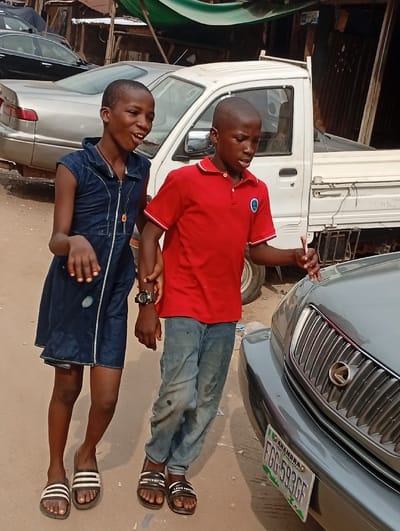 Two children, a girl and a boy, are walking along a street. The girl wears a denim dress and striped sandals, while the boy sports a red polo shirt, jeans, and branded slides. They are holding hands and appear to be in conversation. Cars are visible in the background. The scene takes place on a dusty street. A license plate reading "FGG 593 GF" is visible on a nearby car.