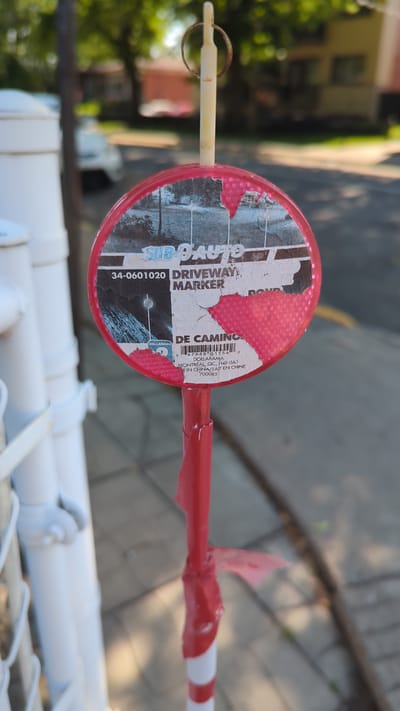 A red and white striped pole with a circular marker is visible in the foreground, positioned on a sidewalk next to a white fence. The marker, which appears to be a driveway marker, has a torn label that reads "SUB O AUTO DRIVEWAY MARKER." Below this, a barcode is visible along with text indicating "DOLLARAMA MONTRÉAL, QC, H4P 0A1, MADE IN CHINA/FAIT EN CHINE 700085." The background is blurred but shows a residential street with houses and trees. The lighting suggests it is daytime.