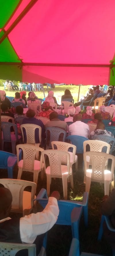 The image shows a gathering of people seated on plastic chairs under a canopy tent in Malava, Kenya. The canopy is red and green. There are many people, mostly women, wearing traditional clothing. The people are seated in rows, facing a grassy area beyond the tent. They appear to be attending a social event, such as a funeral or a wedding. The event is taking place on a grassy field. The location is in the rural area of Malava, Kenya, specifically at coordinates 0.521 latitude and 34.838 longitude.
