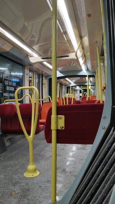 The interior of a bus or tram features rows of red and orange seats with yellow support poles. A yellow, heart-shaped handrail stands near the front. A window is visible, showing what appears to be rain. The floor is speckled gray. Overhead lights illuminate the scene. The accordion-style barrier of a doorway is visible in the right foreground.