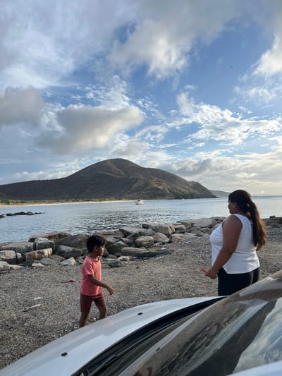 Two people, a young boy and an adult woman, stand on a rocky shoreline near a body of water. The boy, wearing a pink t-shirt and brown shorts, is walking away from the camera. The woman, wearing a white tank top and black pants, is looking out at the water. In the background, a large, dark mountain dominates the landscape. A white boat is visible on the water in the distance. The sky is blue with scattered clouds. The scene appears to be located in the city of Pedro González, Venezuela.