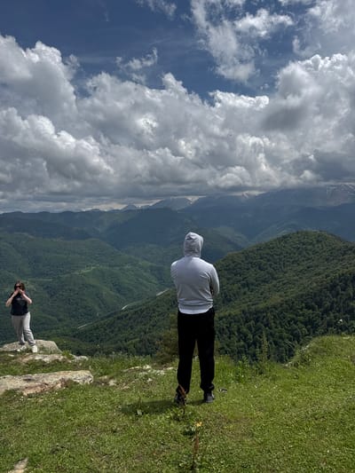 Two individuals stand on a grassy plateau overlooking a vast, mountainous landscape. The person on the left, a woman, is positioned on a rocky outcrop, wearing a dark t-shirt and light-colored pants, and appears to be holding a camera or phone up to her face. The person on the right, a man, stands further back and to the right, wearing a grey hooded sweatshirt and dark pants, with his back to the camera, gazing at the expansive view. The background is dominated by layers of densely forested mountains, receding into the distance, with snow-capped peaks visible on the horizon. The sky is filled with dramatic, volumetric clouds against a blue backdrop. The scene suggests a location in or near Nizhnyaya Teberda, Russia, known for its mountainous terrain and natural beauty.