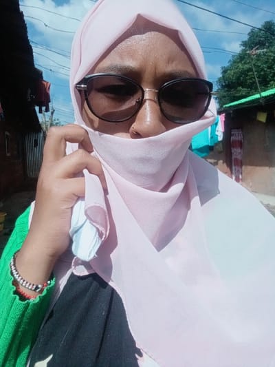The image, taken in Nyeri, Kenya, captures a close-up of an individual, likely female, from the chest up, positioned outdoors under a blue sky with scattered clouds. The person wears a light pink headscarf (hijab) that covers the head and is drawn up to obscure the mouth and chin. Dark-lensed sunglasses with a gold-colored bridge cover the eyes, and a small gold-colored stud is visible in the right nostril. The right hand, with a dark skin tone, is raised, holding the pink fabric near the chin. Multiple beaded bracelets in red, black, and white are worn on the right wrist, over a visible green ribbed long-sleeved garment. A dark garment with light patterns is partially visible beneath the pink fabric.

The background shows an outdoor residential setting. To the left, a structure constructed with corrugated metal siding is visible, with dark clothing hanging to dry. Overhead, multiple black utility wires are strung across the frame. To the right, a building with unpainted light brown walls and a green corrugated iron roof is present, with various colored clothing items hanging outside. Green foliage and trees are visible above this structure.