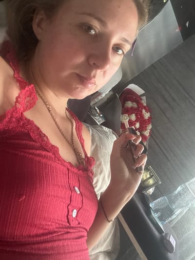 In this image, a young woman with light brown hair is captured from the chest up, lying down. She is wearing a red, ribbed tank top with lace trim and two white buttons. Around her neck, she has a gold chain necklace. Her right hand is raised, holding a small, dark object, possibly a cigarette or vape pen, with neatly manicured nails. In the background, to her right, is a bouquet of red and white artificial roses. The setting appears to be indoors, with a plain wall in the background and what looks like a bed or soft surface she is lying on. There are also some bottles and other objects visible on a surface behind the bouquet. The overall lighting suggests natural light coming from the side.