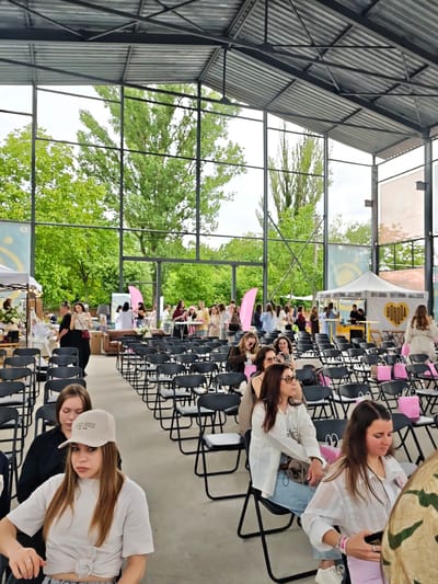 This image depicts a daytime event taking place within a large, open-sided structure with a corrugated metal roof supported by exposed black metal beams and trusses. The background features a panoramic view of lush green trees and a bright, overcast sky. The event is located in Vinnytsia, Ukraine, identified by a white canopy tent in the background displaying a yellow heart logo with the word "BORODA."
In the foreground, numerous black folding chairs are arranged in rows on a concrete floor. On the left, a woman with light brown hair wears a white baseball cap and a white t-shirt tied at the waist, looking towards the viewer. A silver ring is visible on her left ring finger. Behind her, another woman with light brown hair wears a black long-sleeved top and looks downwards. On the right, a woman with dark hair wears a white textured long-sleeved shirt and looks down at a handheld electronic device. A pink paper bag is visible next to her. Further right, a woman with dark hair and glasses, dressed in a white collared shirt and light blue jeans, is seated, looking towards the left.
The mid-ground contains additional rows of black folding chairs, many occupied by women. Several pink paper bags are placed on or adjacent to the chairs throughout the seated area.
The background displays active event stalls and groups of individuals. On the left, a white canopy tent serves as a vendor booth with various items displayed and a woman in a black shirt behind a counter. Towards the right, the aforementioned white canopy tent with the "BORODA" logo shows two men behind a counter, appearing to serve customers. A woman in a light-colored long coat is walking away from this tent. In the deeper background, a brown leather sofa, additional tables, and smaller groups of people are visible, indicating areas for socializing or product displays. Large fabric banners with abstract designs, including a blue banner with a white star-like emblem on the left, are affixed to the structural supports. The event appears to be a gathering or market with both a formal seating area and informal vendor zones.