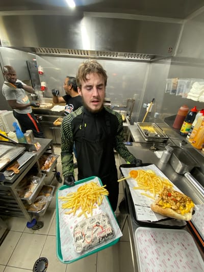 Inside a commercial kitchen, a fair-skinned person holds trays of fries and sandwiches. Two other workers are present. One is slicing food. The kitchen features stainless steel surfaces, fryers, and condiment bottles. Trays are lined with paper, displaying fries and sandwiches. The scene is brightly lit with fluorescent lighting.