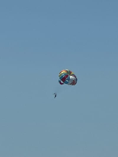 A multi-colored parachute is descending in the sky, with a person attached below. The parachute is round and appears to be made of fabric, with sections of red, yellow, blue, and white. The person is small in comparison to the parachute and is in a freefall position. The sky is a clear, pale blue. The location is the city of Boreti, Montenegro.