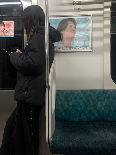 The image depicts the interior of a commuter train car in Nakano, Japan. A solitary individual, facing away from the camera, occupies the left side of the frame, leaning against a vertical silver handrail. This person has dark, shoulder-length hair with lighter streaks and is dressed in a black puffer jacket with a hood, paired with a black long skirt or wide-leg pants featuring silver stud embellishments down the side. Both hands hold a black smartphone, with thumbs positioned for active interaction. The train interior features white walls, a light gray ceiling, and green patterned seating with a checkerboard design on the seatback and a textured pattern on the cushion. Two advertisements are visible: a partial "CEZANNE" cosmetic ad with a smiling woman on the left, and a larger ad on the right displaying a male model against a light blue background, advertising "ジョブメドレー" (Job Medley) and claiming "求人数No.1" (Number 1 in job openings). A small etiquette sign in Japanese is positioned above the Job Medley advertisement.