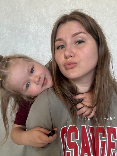 The image shows a woman and a young girl, likely a mother and daughter. They are positioned close together, with the girl slightly below the woman. Both have fair skin and light eyes. The woman has brown hair and is wearing a grey shirt with red and white lettering; the lettering appears to read "CHECK STARS". The girl has blonde hair pulled back in small pigtails. The background is a plain, light-colored wall. The photograph appears to have been taken indoors. There is no indication of the location being in the city of Gornyi (Dvurechenskoe S/P), Russia, based on the visible information.