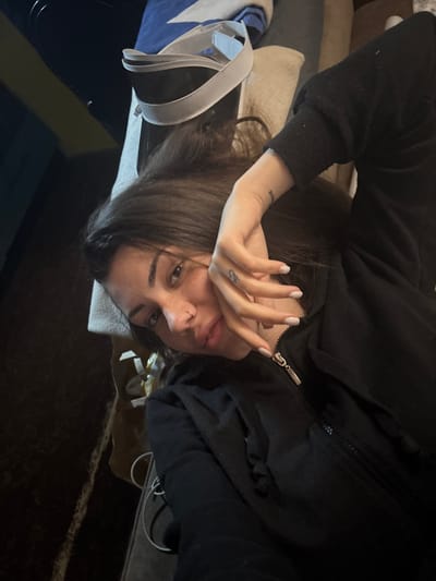 An indoor photograph, likely taken in a casual setting in Belopoptsi, Bulgaria, features a young woman lying down or lounging, looking directly towards the camera. She has long dark hair and is wearing a black zippered hoodie or jacket. Her left hand is raised, partially covering the lower half of her face, with her fingers gently spread. Her fingernails are painted a light, neutral color, possibly white or light pink, and each features a small, subtle embellishment. A small, dark tattoo is visible on the back of her left hand, between her thumb and index finger, and another faint tattoo can be seen on her left wrist/forearm. She has a small ring piercing in her right nostril. Her lips are slightly parted. The room is dimly lit, suggesting it is either evening or nighttime. The background consists of dark, soft furnishings, possibly a couch or bed, with glimpses of other objects including a white strap, a blue and white item, and a white coiled cable. The overall mood is relaxed and informal. No discernible text is visible in the image.