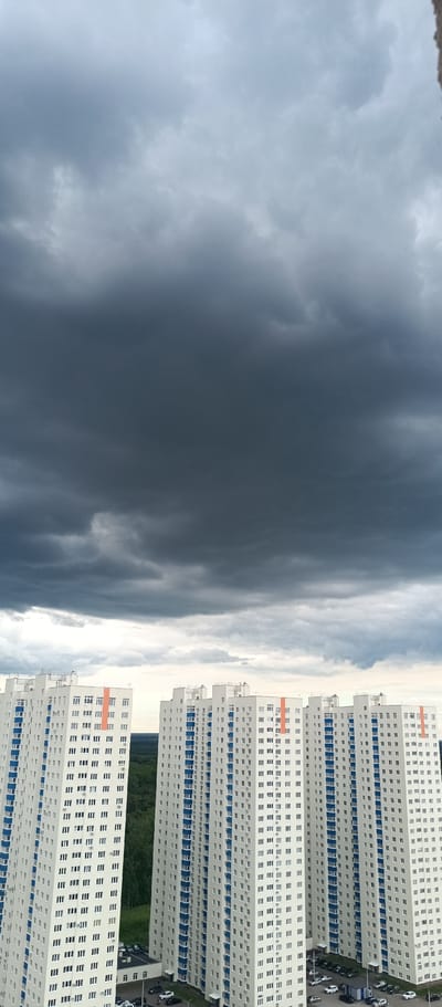 The media file depicts a dramatic sky filled with dark, ominous storm clouds that dominate the upper two-thirds of the frame. Below the brooding sky, two identical, modern apartment buildings stand tall, their white facades adorned with vertical blue accents and orange stripes. The buildings are positioned close together, creating a sense of urban density. In the foreground, cars are parked in a lot at the base of the buildings, and a line of green trees is visible between the structures.
The lighting suggests it is daytime, likely late afternoon or early evening, given the heavy cloud cover and the contrast between the bright buildings and the dark sky. The weather appears to be inclement, with the dark clouds indicating an approaching storm or the remnants of one. The scene is likely set in a residential area of Ufa, Russia, given the architectural style of the buildings and the presence of the dense forest in the background, which is common in the outskirts of cities. The overall mood is somber and dramatic, evoking a sense of impending weather change. There is no visible text in the image.