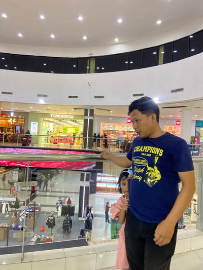 In an indoor shopping mall, a man in a dark blue t-shirt with a "Champions NBA 2015 Golden State Warriors Steph Curry" graphic stands at a railing. He is looking to his left. Partially obscured behind him, a young girl in a pink dress peeks out, also looking towards his left. The man's left hand rests on the railing. The background shows multiple levels of the mall with storefronts, including one labeled "TOMORROW." The environment is well-lit with overhead lights and displays. The scene is consistent with a shopping mall in Sidoarjo, Indonesia.