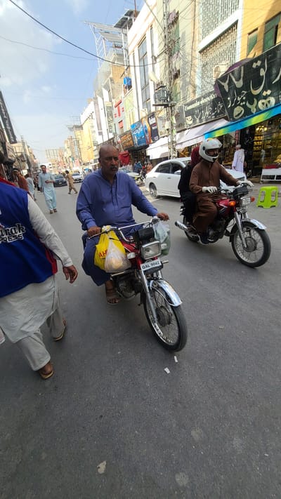 Here is a detailed description of the image:

**Scene:**
The image appears to be taken on a busy street in Quetta City Tehsil, Pakistan. The street is lined with buildings and various commercial establishments with signage in Urdu and English.

**People:**
*   A man is sitting on a red motorcycle, holding a yellow plastic bag with a loaf inside. He wears a blue tunic.
*   Another man is riding a motorcycle wearing a brown outfit and a white helmet.
*   A man is lying on the street wearing traditional shalwar kameez.
*   Several other people are seen walking on the street in the background, mostly wearing traditional Pakistani clothing.

**Objects:**
*   Two motorcycles are prominent, one red and one brown.
*   A yellow plastic bag with a loaf inside.
*   Various storefront signs with text including "JEWLERS" and "PRESS".
*   A white car is parked on the side of the road.

**Environment:**
*   The image is taken in daylight.
*   The street is paved and shows signs of wear and tear.
*   The buildings have a mix of old and new architectural styles.

**Location Clues:**
The presence of signage in Urdu and the clothing styles suggest a South Asian location. The license plate on the red motorcycle says "QAL 9042" which corresponds to vehicles in the Quetta district.