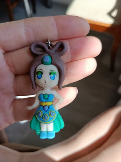 A hand is holding a small, colorful figurine keychain. The figurine depicts a young girl with brown hair styled in a bun, large blue eyes with green irises, and a light-colored face. She is wearing a blue and yellow dress with green trim and blue shoes. A small green gem is visible on her chest and another on her forehead. The keychain loop is attached to the top of her head. The background is blurred, with hints of sunlight and furniture. The image does not provide any information about the city of Odesa, Ukraine.