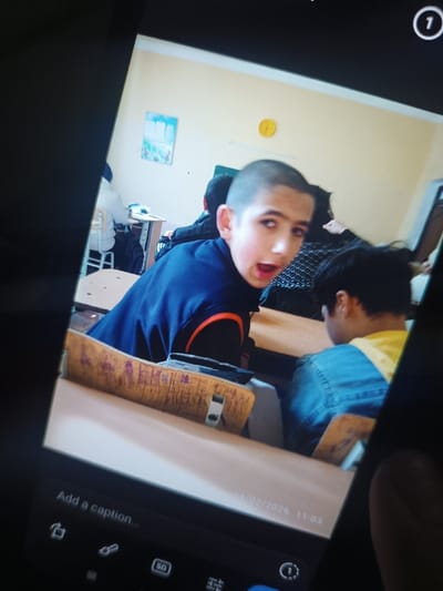 A student with a shaved head and a surprised expression peers over his shoulder in a classroom. He is wearing a blue and orange jacket and sitting at a desk. Other students and desks are visible in the background. The photo is taken of a screen, showing an image captured on February 14, 2026. A caption bar and various icons are visible at the bottom of the screen.