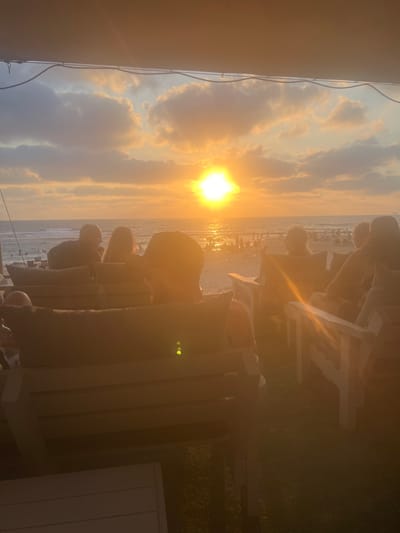 The image captures a serene sunset scene at a beach, likely in Hof HaCarmel Regional Council, Israel. The sun, a brilliant orb, casts a warm, golden glow across the sky, illuminating scattered clouds. The light reflects intensely off the water, creating a shimmering effect. In the foreground, several individuals are seated in what appear to be Adirondack chairs, facing the ocean and the sunset. Their postures suggest relaxation and appreciation of the moment. Some are silhouetted against the bright sky, while others are more visible, their features softened by the low light.
The setting is a grassy area adjacent to the beach, with several white chairs providing comfortable seating. The presence of lounge chairs and the view of the ocean indicate a beachside resort, cafe, or private residence. The overall atmosphere is peaceful and tranquil, suggesting a moment of quiet contemplation or shared enjoyment of the natural beauty. The time of day is clearly sunset, given the intense golden light and the low angle of the sun. The weather appears to be clear, with only scattered clouds adding texture to the sky. There is no visible text in the image.