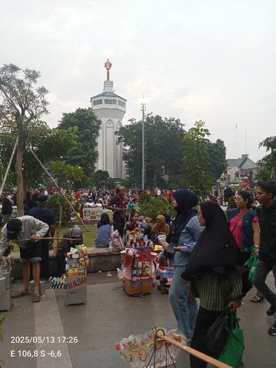 The image depicts a bustling street scene in the city of Bogor, Indonesia (latitude: -6.595, longitude: 106.792). Numerous people are gathered in a park-like setting, some sitting on benches and some standing around. Several vendors are selling snacks and beverages from mobile carts. Several people are interacting with each other, while others are browsing the vendors' goods. In the background, a tall, white tower with a gold spire stands out, marking the location. There are various trees and bushes surrounding the area, providing a lush backdrop. The sky is cloudy and overcast. The image captures a typical everyday scene in the city of Bogor, Indonesia.