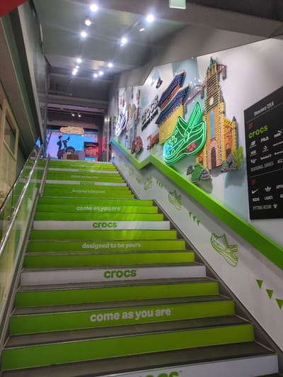 This image captures an interior view of an upward-sloping staircase within a commercial establishment, identified as a Crocs location in Myeongdong, Seoul, South Korea. The staircase consists of steps alternately colored green and white, with the green risers featuring Crocs brand slogans such as "crocs™", "come as you are™", and "designed to be yours" in white text. A transparent glass railing with a metallic top handrail ascends along the left side of the staircase.
On the right wall, a large, multicolored 3D art installation depicts various architectural structures, including a traditional Korean building and a European-style church, both seemingly constructed from small, distinct objects. A prominent neon green outline of a Crocs clog is integrated into this artwork, along with the words "crocs" and "MYEONGDONG". Adjacent to this art, a black directory board, labeled "Directory 안내", indicates "2F" (Second Floor) houses "crocs", "new balance", "FILA", "PUMA", "Vans", "Timberland", "SKECHERS", "Converse", "Dr. Martens", "Stans", "SEASONAL SHOES", and "ACC" (Accessories). "1F" (First Floor) lists "NIKE", "ADIDAS", "APPAREL", "KIDS", "LADIES", and a "FITTING ROOM". The lower section of the right wall along the staircase displays green outlines of Crocs clogs and triangular motifs.
At the top of the stairs, a large video screen shows a male individual in a plaid skirt and jacket, striking a pose or dancing, beneath a graphic bubble containing the text "do your thing". A small display stand bearing the Crocs logo and the phrase "do your thing" is positioned in front of the screen. The ceiling is grey and equipped with multiple bright, recessed spotlights illuminating the scene.