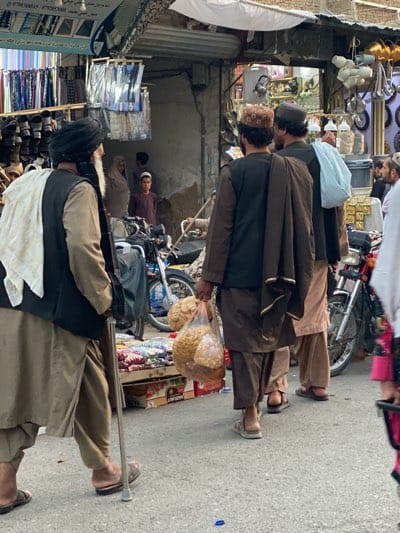 The image depicts a bustling street scene in Kandahar, Afghanistan. Several men are walking along a paved road adjacent to shops. The man in the foreground is older, with a white turban and a black vest over a tan robe; he uses a cane. Another man carries a plastic bag filled with food. In the background, motorcycles are parked near open storefronts displaying merchandise, including shoes and clothing. Other men and a boy are visible in the background.