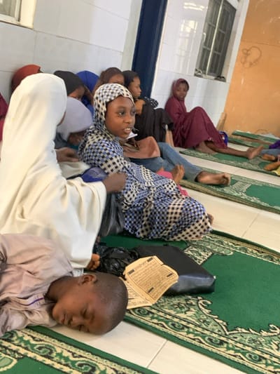 A group of people are lying on a floor. They appear to be resting or sleeping. Some are on a green rug with patterned designs. A woman in a blue and white checkered headscarf is lying on her side looking up. A young boy in a light brown shirt is lying on the rug with his eyes closed. There is a black bag and some papers nearby on the rug.