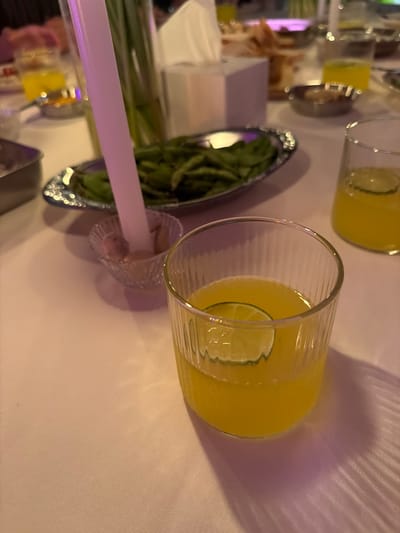 A dimly lit, formal dining scene is depicted, likely in Chang Phueak, Thailand, with a white tablecloth covering the table. In the foreground, a clear glass filled with a bright yellow beverage and a slice of lime is prominently featured, its textured surface catching the ambient light. To the right, another glass of the same yellow drink is partially visible. In the mid-ground, a silver serving dish holds a pile of green edamame, and a white candle stands upright in a small, crystal dish containing a light-colored substance, possibly salt. Behind the edamame, a white tissue box and other food items on silver platters are scattered across the table. The lighting is soft and diffused, with hints of purple and pink hues, creating a warm and intimate atmosphere.