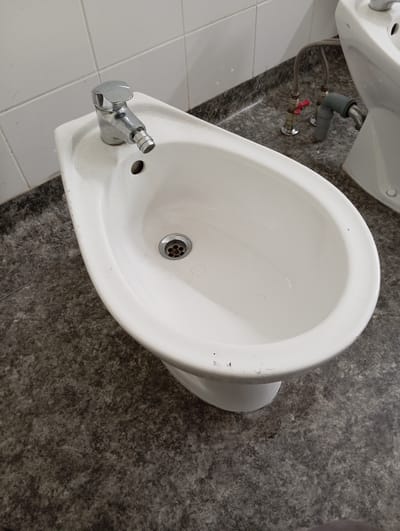 The image shows a bidet in a bathroom. The bidet is white ceramic with a chrome faucet. The faucet has a single handle and a bidet spray attachment. The floor is tiled with a dark gray speckled pattern. In the background, a toilet is partially visible. Water supply lines and a drainpipe are also visible behind the bidet. The scene appears to be in a residential or public restroom.
