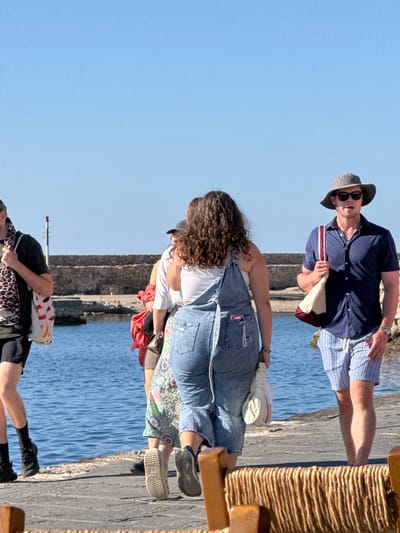 In Chania, Greece, on a sunny day, a group of four individuals walks along a stone-paved promenade next to the clear blue sea. The person in the foreground, on the right, is a man wearing a navy blue short-sleeved collared shirt, striped blue and white shorts, a wide-brimmed grey hat, and sunglasses. He carries a cream and burgundy canvas bag. To his left, a woman with curly brown hair is visible from the back, wearing denim overalls over a white top and carrying a cream-colored bag. Behind her, another woman is partially visible, wearing a floral patterned dress. Further to the left, a person wearing a black shirt and shorts, a leopard-print scarf, and carrying a multi-colored tote bag, walks away from the camera. In the background, a stone wall and a red and white lighthouse structure line the waterfront.