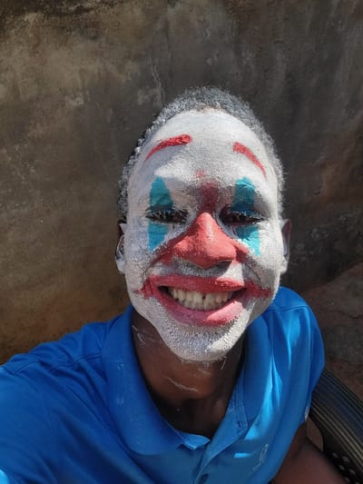 A person is lying on a concrete surface in an outdoor setting, with the head oriented towards the top of the frame. The individual is wearing a white clown mask with painted red details around the mouth and on the nose, and blue markings around the eyes. The mask appears to be cracked and textured. The person is wearing a blue shirt. The setting appears to be in Nkpor, Nigeria, as specified in the prompt.