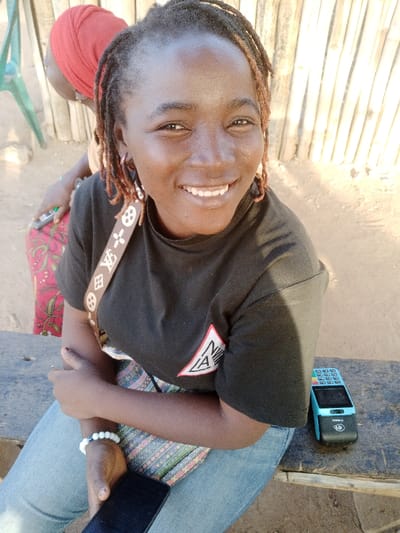 A smiling woman with dark hair and a black t-shirt sits outdoors. She wears light blue jeans, a bracelet, and earrings. A patterned handbag strap crosses her chest. Next to her is a dark phone and a blue and black point-of-sale device. Behind her, a person in a red head covering sits near a light green chair with a bamboo fence in the background. The scene is lit with natural daylight.