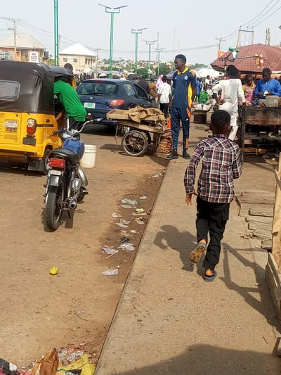 This image depicts a street scene in Tempure, Nigeria. In the foreground, a young boy wearing a plaid shirt and black pants runs away from the camera on a concrete walkway. He is wearing blue sandals. To the left, a yellow auto-rickshaw is parked on a dirt road. A person in a green shirt is partially visible inside the rickshaw. A blue motorcycle is parked behind the rickshaw. The background shows several buildings, including what appears to be a mosque and other shops. There are also people walking and standing around, some near food stalls. The environment appears to be a busy marketplace or roadside area with some scattered litter on the ground. Streetlights are visible in the distance.