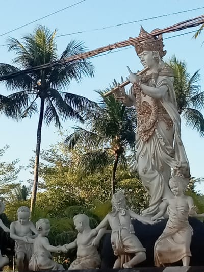 A statue depicting the Hindu deity Krishna playing a flute stands prominently in Blahbatuh, Indonesia. Krishna is adorned with a crown and ornate jewelry, with flowing garments. He is surrounded by smaller statues of children, who appear to be dancing in a circle. The statues are light-colored, possibly made of stone or plaster. In the background, lush green palm trees and other foliage are visible, under a clear blue sky. Overhead, power lines are strung across the scene. The overall impression is one of a religious or cultural monument situated in a tropical environment.