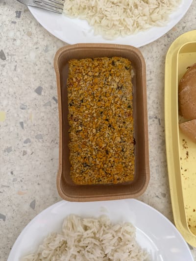 A top-down view shows a meal laid out on a speckled grey countertop, likely in Milan, Italy, as suggested by the context of the image. In the center is a rectangular brown paper tray holding a breaded food item, possibly chicken or fish, covered in coarse golden-brown breadcrumbs with flecks of green herbs and red spices. To the left of the tray, a portion of white rice is visible on a white plate, next to a metal fork. To the right of the tray is a yellow plastic tray containing two halves of a bread roll. Another white plate with white rice is positioned in the lower part of the image. The lighting is bright and even, suggesting an indoor setting. No people are visible in the image, and there are no specific architectural features to identify the exact location within Milan. The overall scene depicts a simple, prepared meal.