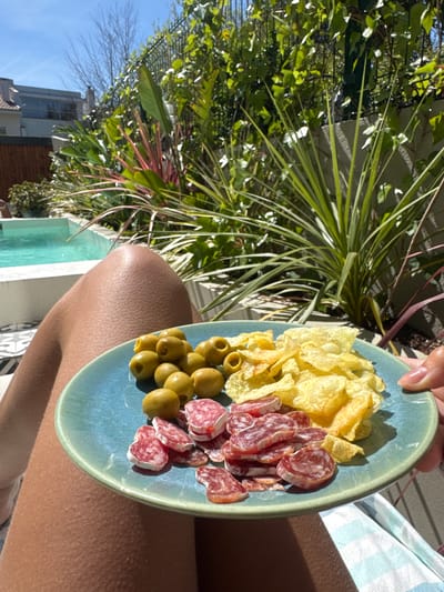 A person holds a light blue plate with potato chips, green olives, and sliced sausage in the vicinity of an outdoor area with green foliage, including a yucca-like plant with long leaves. In the background are a building and a blue sky. The person is wearing light-colored clothing. The location is possibly in Porto, Portugal, judging by the vegetation, architecture, and light.
