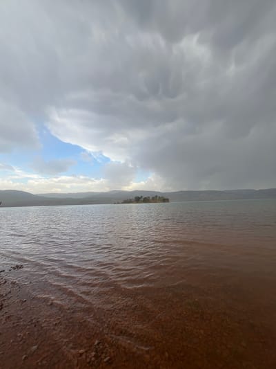 The image, oriented as a landscape, depicts a vast reservoir or lake, likely the Bin El Ouidane Dam in Morocco, set against a backdrop of mountains under a cloudy sky.
In the immediate foreground, a reddish-brown, pebbly shoreline meets the water. The water itself appears calm but textured with ripples, reflecting the diffused light from the sky. Its color is a muted brownish-gray. Across the expanse of water, a narrow strip of land in the midground features scattered green vegetation and a cluster of small trees. Beyond this, a range of rugged mountains stretches across the horizon, appearing hazy in the distance.
The sky occupies a significant portion of the image, dominated by heavy, dark gray clouds that suggest an overcast or impending weather change. However, a distinct area towards the left reveals brighter, whiter clouds and a patch of clear blue sky, hinting at the sun trying to break through, possibly during the late afternoon or early morning hours.
No people or specific activities are visible, emphasizing the natural grandeur and serene, yet dramatic, atmosphere of the mountainous lake environment. There is no visible text in the image.