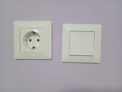 The image shows two white electrical fittings, an electrical outlet and a light switch, mounted side-by-side on a light purple wall in an indoor setting in Roza, Bulgaria. The electrical outlet, located on the left, is a Schuko type, featuring a round receptacle with two main pin holes and top/bottom grounding contacts. To its right, the light switch is rectangular with a single, flat, white rocker button. The brand name "Legrand" is subtly embossed on the lower frame of both the outlet and the switch. The wall has a smooth, painted finish, and the fixtures appear clean and well-maintained. No activity is depicted in the static shot, which focuses solely on the installed electrical components. The lighting is uniform, suggesting an interior environment.